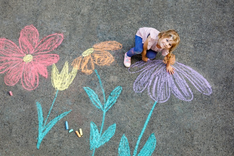 Little preschool girl painting with colorful chalks flowers on ground on backyard. Positive happy toddler child drawing and creating pictures on asphalt. Creative outdoors children activity in summer
